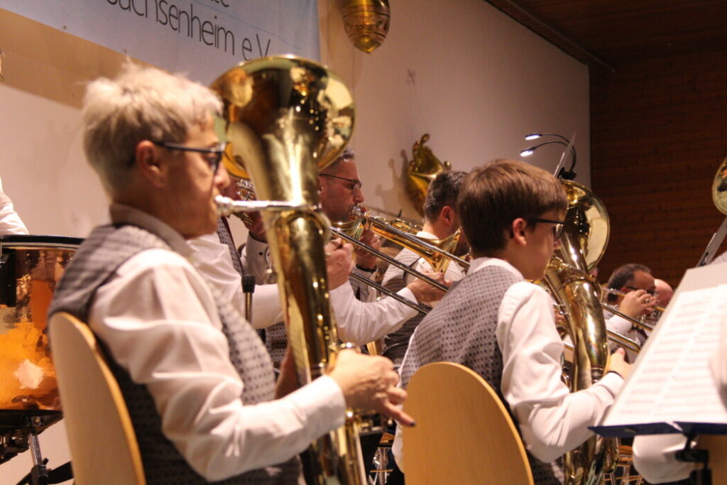 A brass band ensemble is performing indoors. Members are playing instruments such as trumpets and tubas, dressed in white shirts and checkered vests, focusing on their music sheets, creating a formal and coordinated atmosphere.